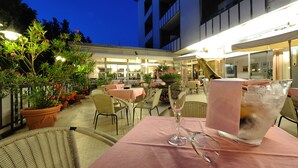 Un área de comedor al aire libre con mesas y sillas, plantas en macetas y un arreglo floral grande.