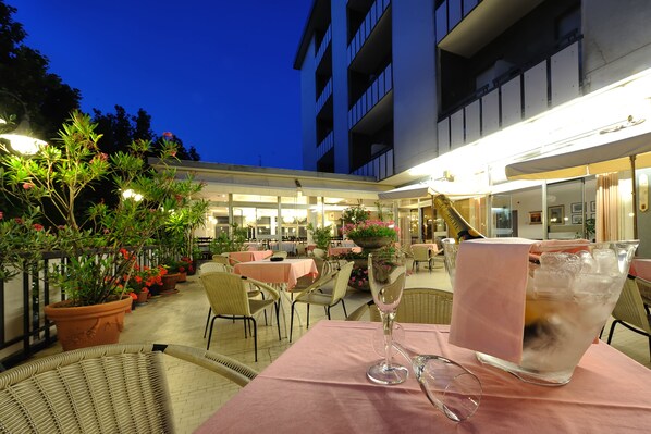 Un área de comedor al aire libre con mesas y sillas, plantas en macetas y un arreglo floral grande.