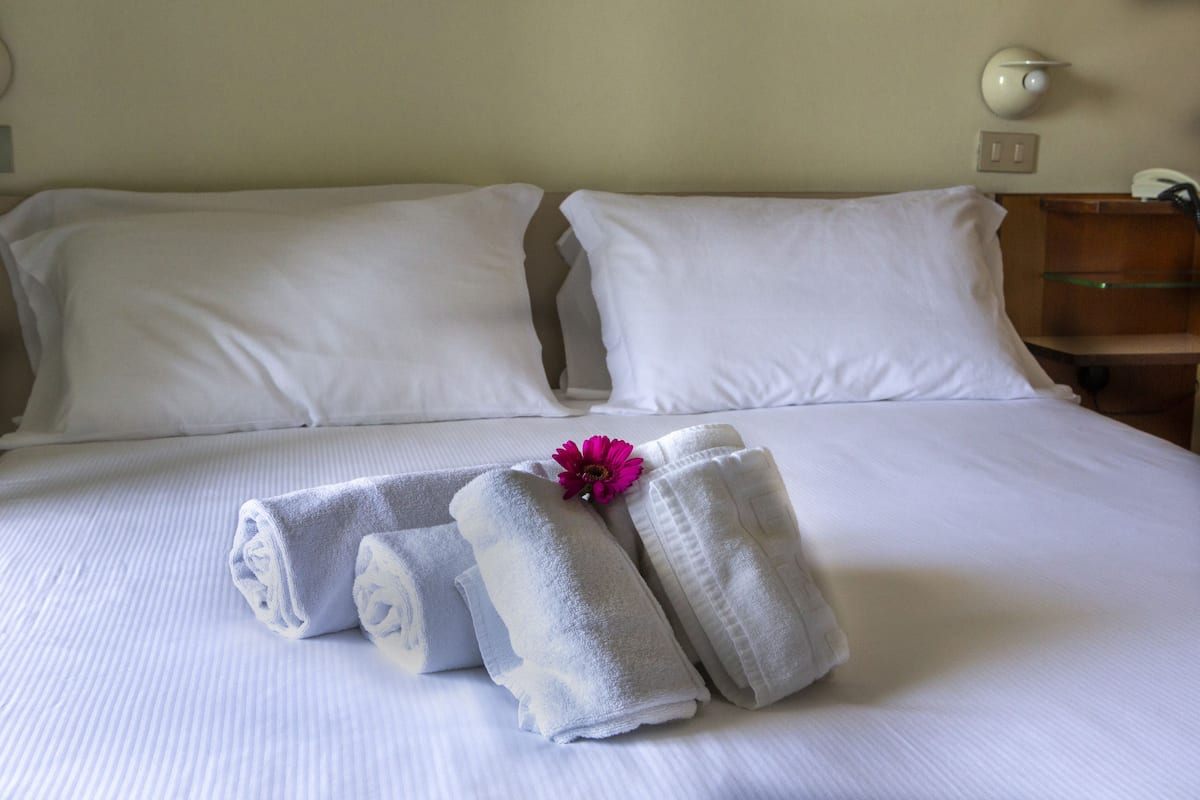A neatly made bed with white linens, a set of towels, and a single flower.