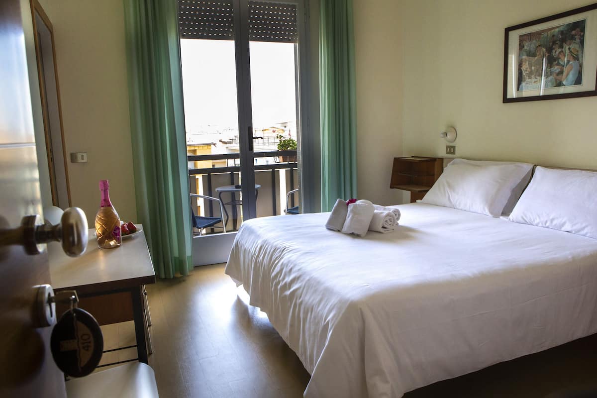 A hotel room with a bed, a table with a bottle and a plate, a chair, a window with curtains, and a painting on the wall.