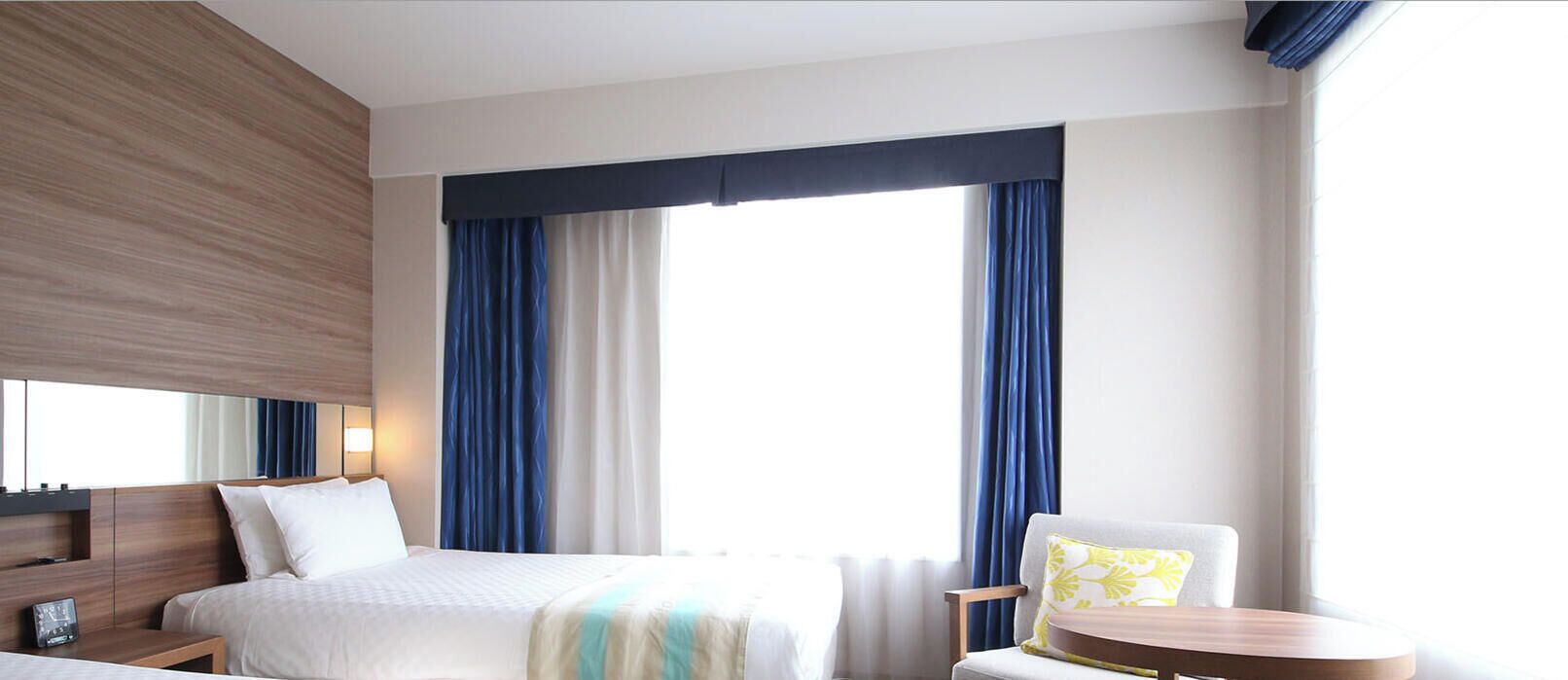 Ein Hotelzimmer mit einem Bett, einem Sessel, einem kleinen Tisch, einem Nachttisch und einem großen Fenster mit Vorhängen.