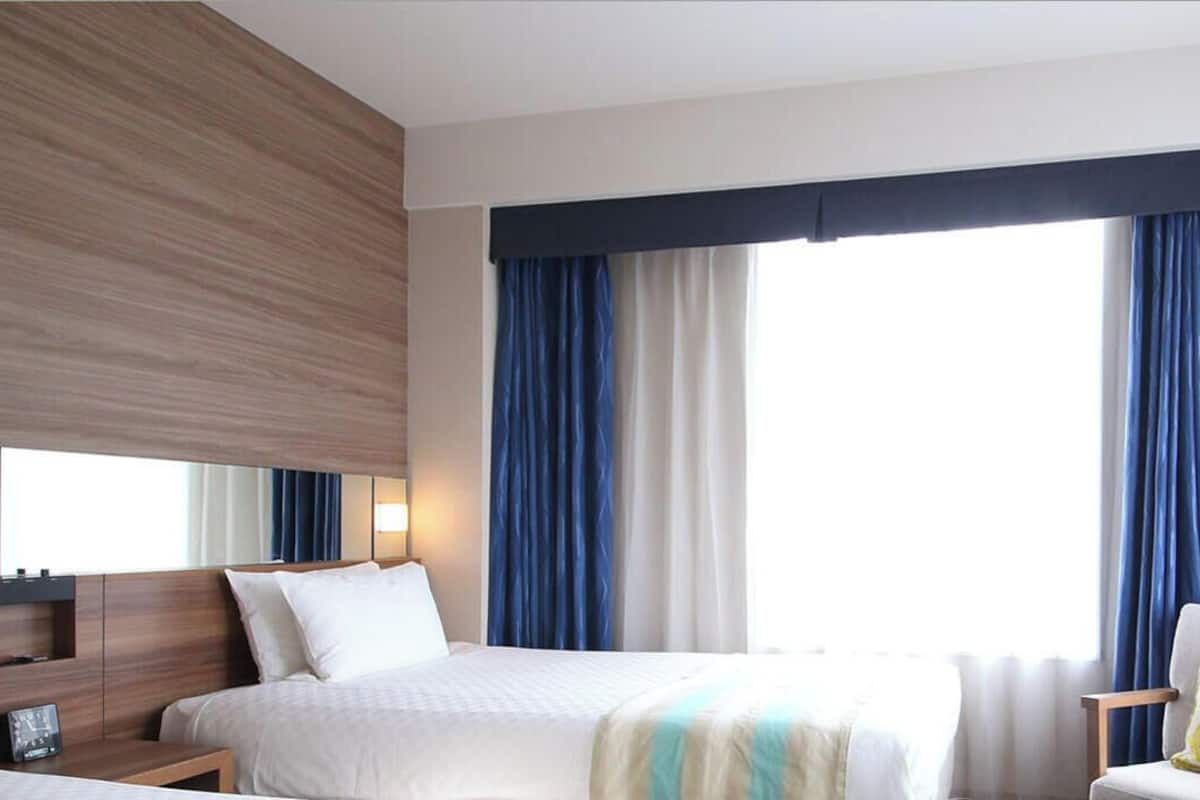 Une chambre d’hôtel avec un lit, une chaise, une petite table, une table de chevet et une grande fenêtre avec des rideaux.