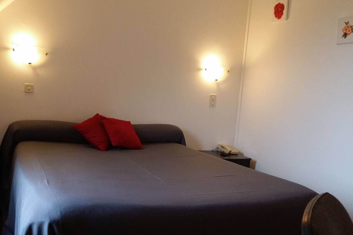 A single bed with a dark bedspread and a red pillow in a room with two wall-mounted light fixtures.