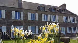 A stone building with multiple windows and shutters, a fenced garden with yellow and white flowers, and a stone bench.