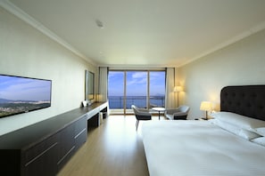A hotel room with a large bed, a TV mounted on the wall, a desk with a chair, a small table with a chair, and a balcony with a view of the sea. - Booyoung Hotel & Resort (Seogwipo)