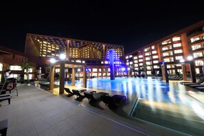 Un patio de hotel con piscina, sillones de descanso y un edificio moderno con ventanas iluminadas.