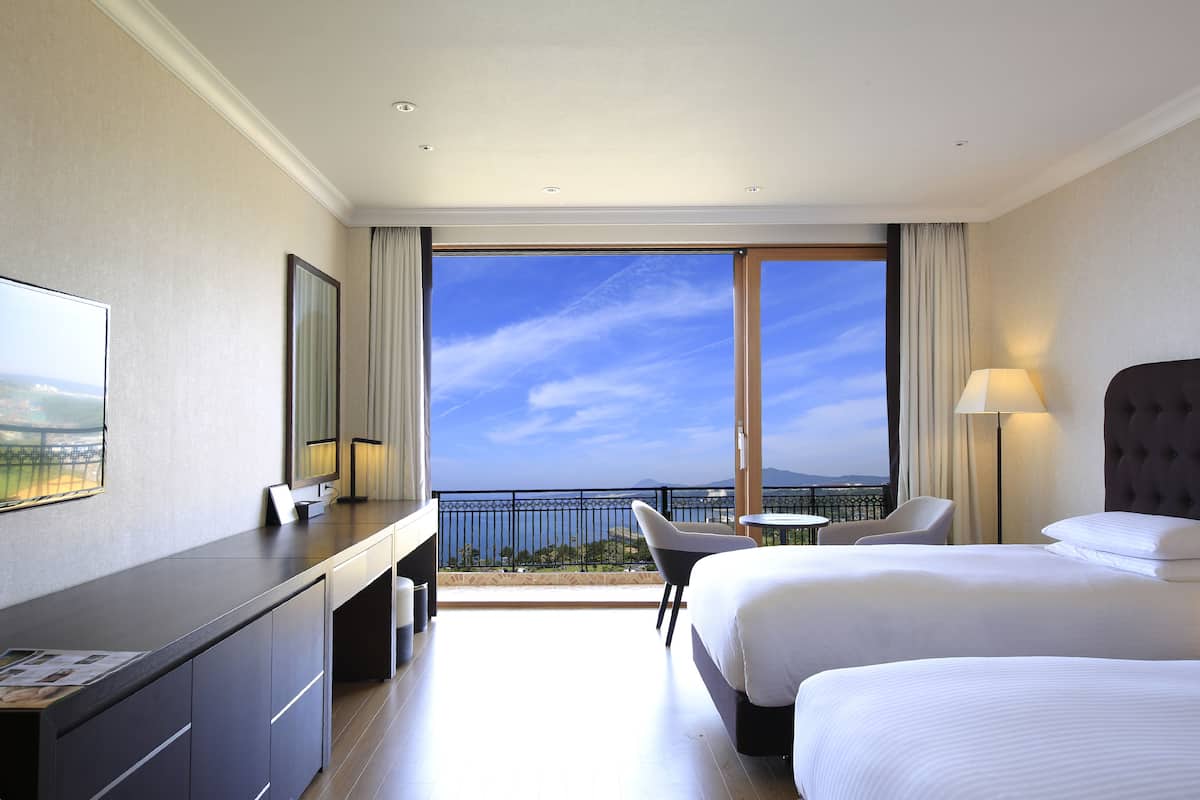 A hotel room with a large window, a desk, two beds, and a chair.