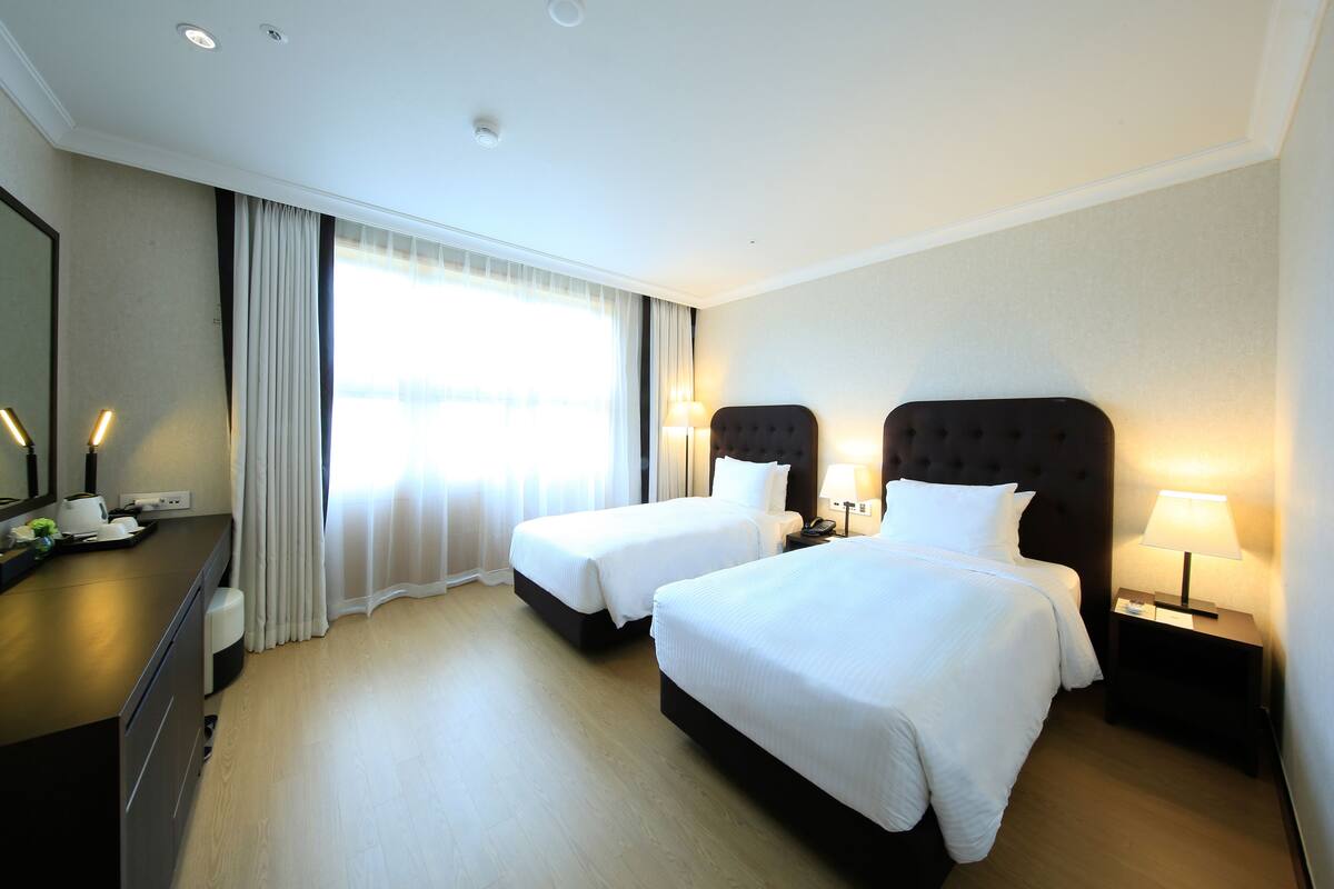 A hotel room with two beds, a desk with a lamp, and a large window with curtains.