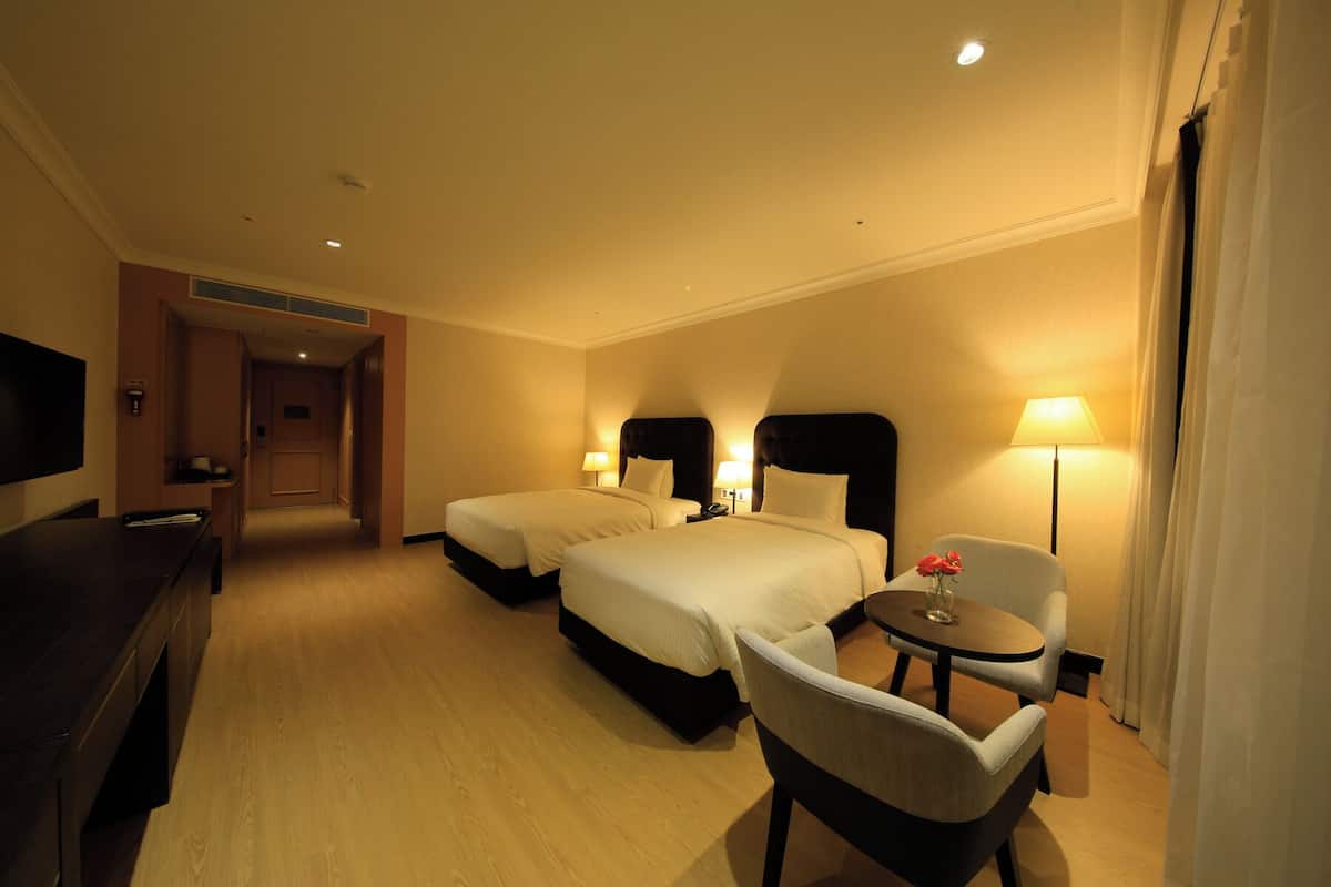 A hotel room with two beds, a desk, a chair, and a small table with a vase of flowers.