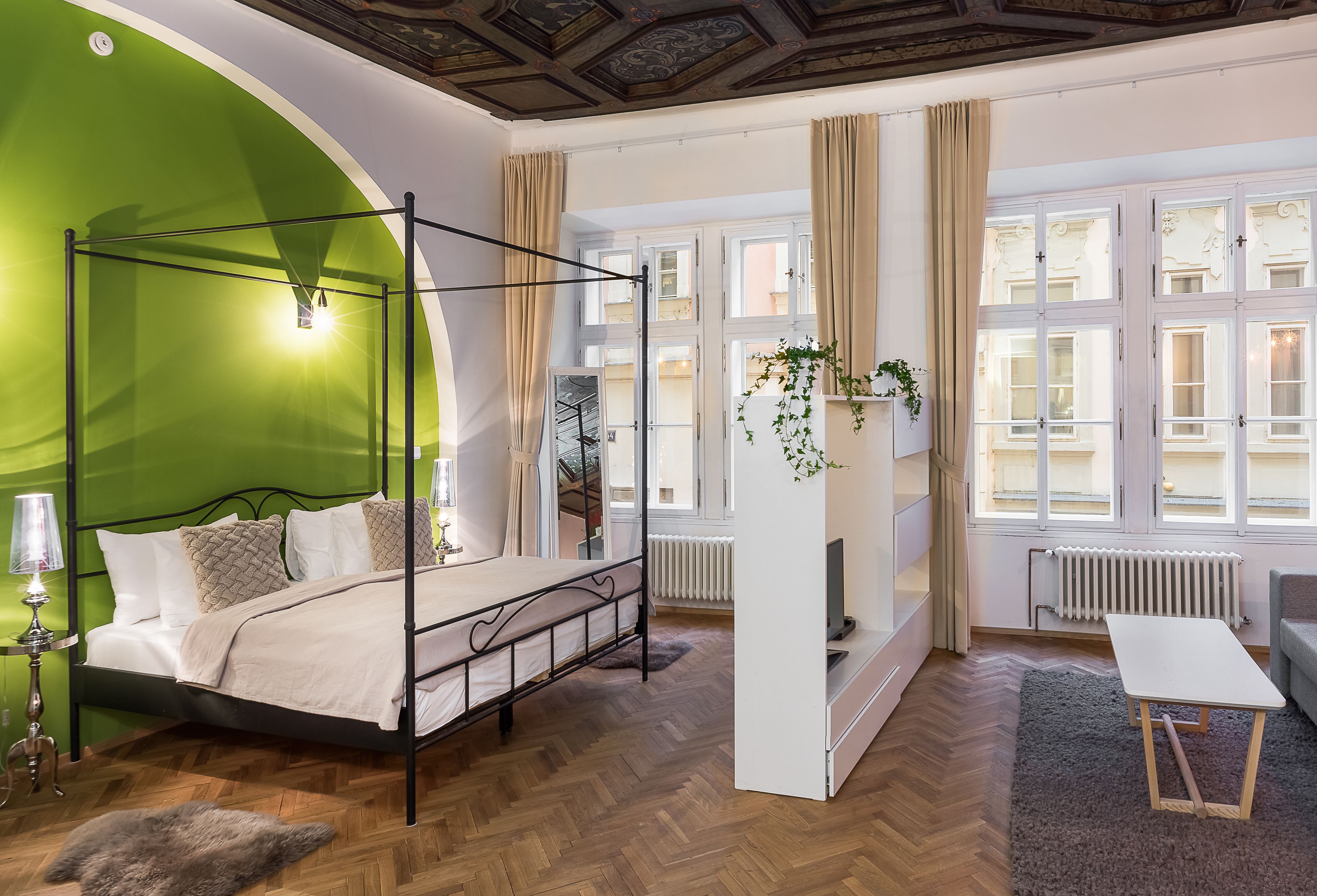 A bedroom with a four-poster bed, a television, a small table, and a view of the city through large windows.