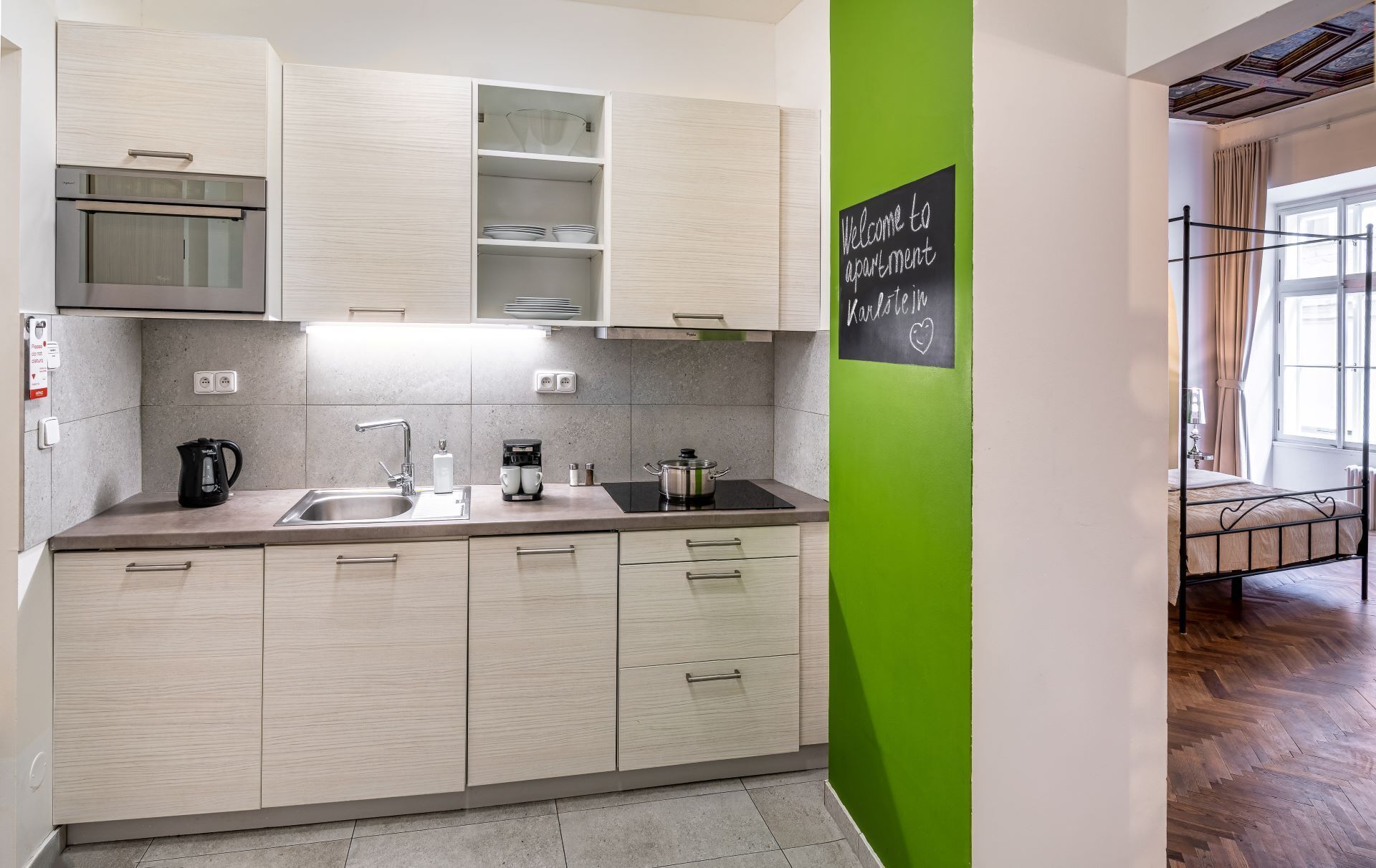 A modern kitchen with light wood cabinets, a stainless steel sink, and a black kettle. There's a chalkboard sign on the wall.
