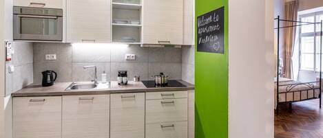 A modern kitchen with light wood cabinets, a stainless steel sink, and a black kettle. There's a chalkboard sign on the wall.