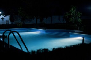 Una piscina iluminada de noche, con zonas oscuras alrededor y algunos árboles.