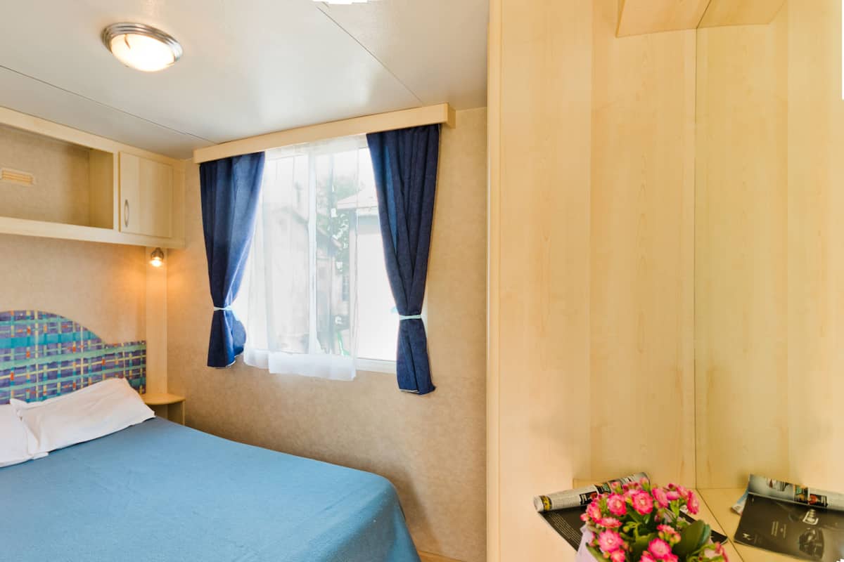 Un dormitorio con una cama, una ventana con cortinas, una mesita de noche, un armario de madera y una pequeña mesa con una revista y flores.