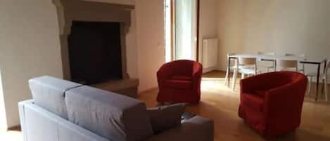 Una sala de estar con un sofá gris, un sillón rojo y una chimenea. Se observa un comedor con mesa y sillas al fondo.