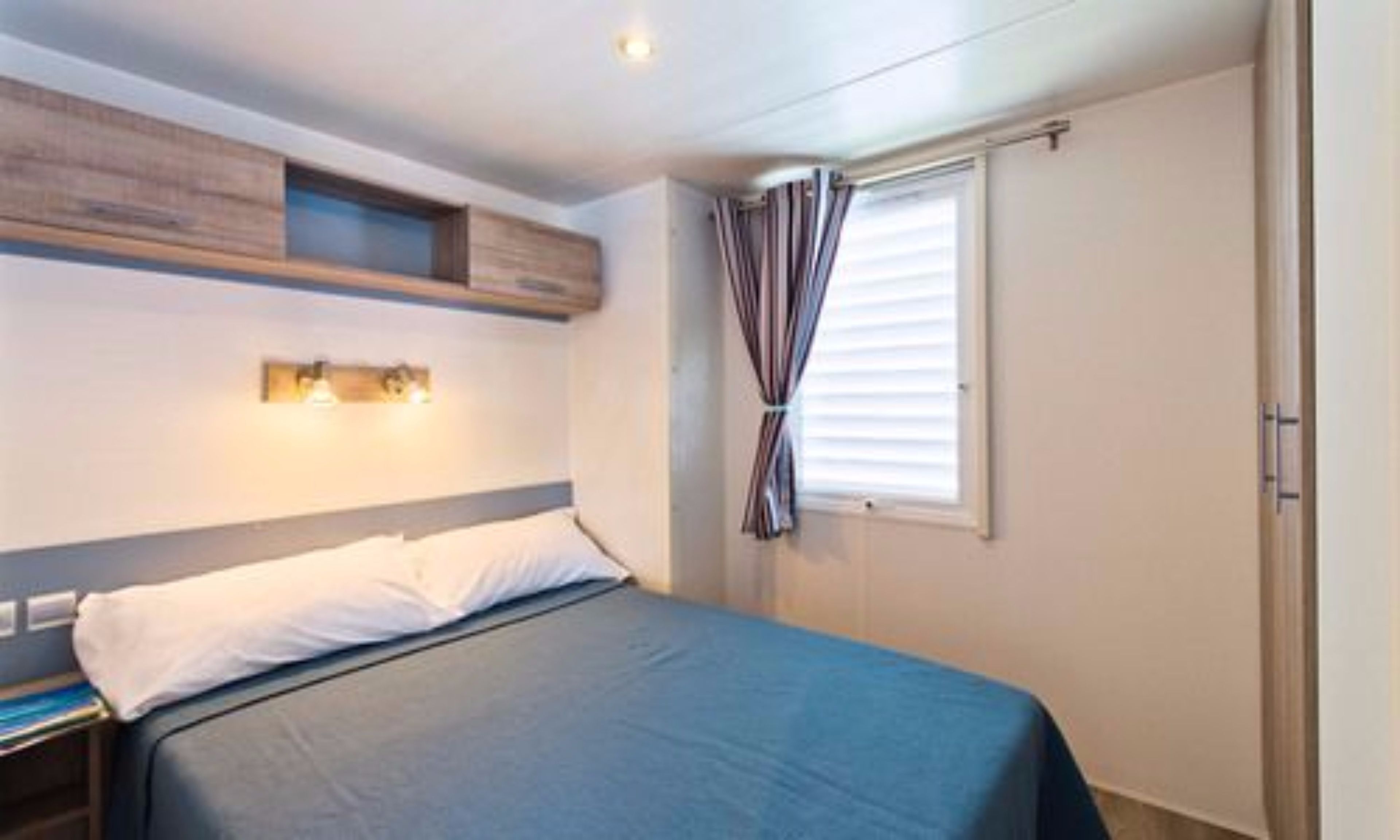Une chambre à coucher comprenant un lit, une table de chevet, une étagère fixée au mur, une fenêtre avec des rideaux et une porte.