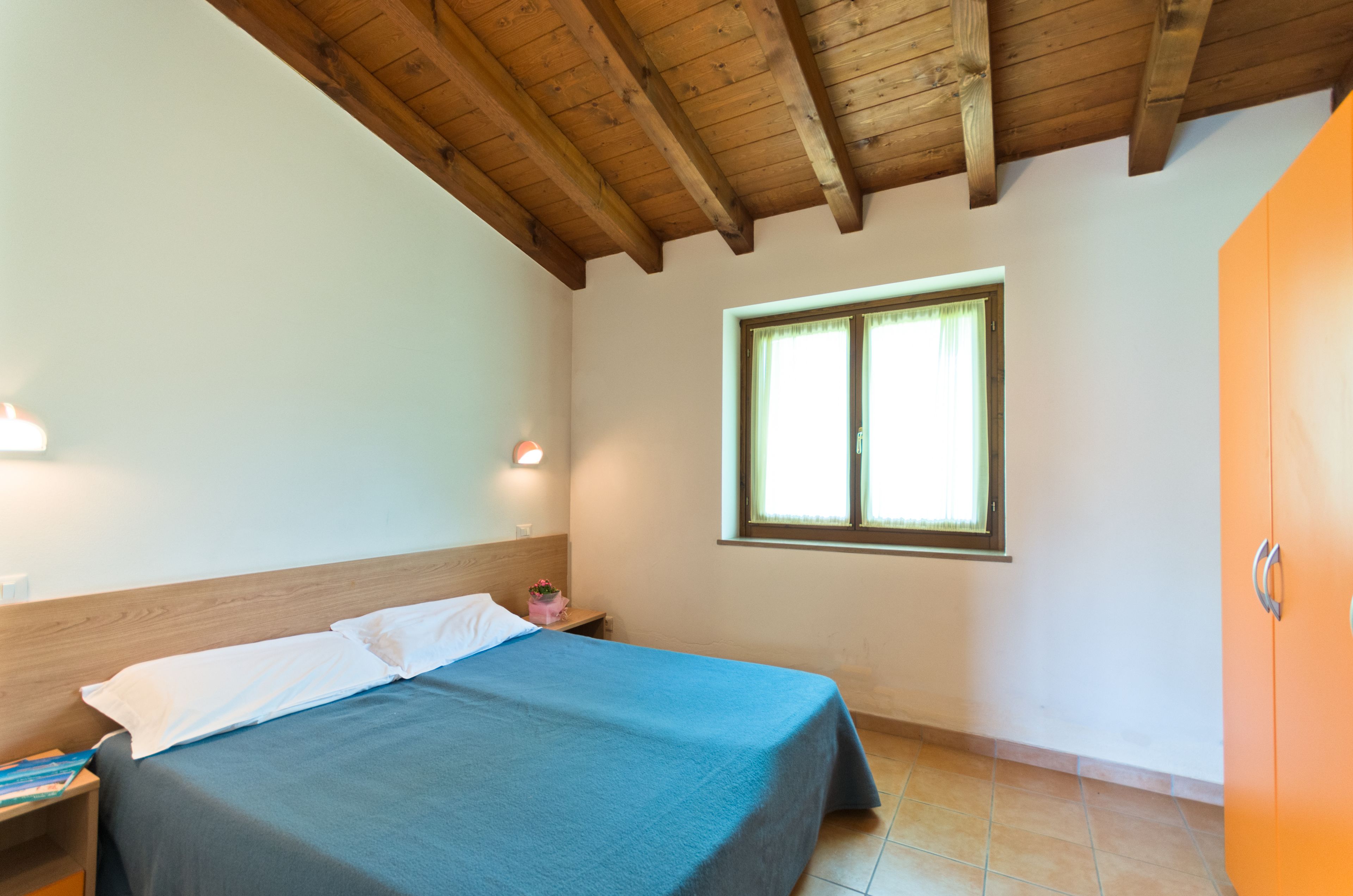 A bedroom with a wooden ceiling, a bed with a blue bedspread, a nightstand with a lamp, a window with blinds, and an orange wardrobe.