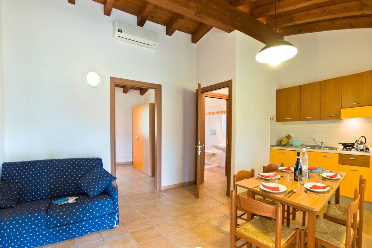 Un espacio habitable compacto con sofá, una mesa de comedor preparada para cuatro personas y una zona de cocina con armarios de madera y un salpicadero de azulejos.