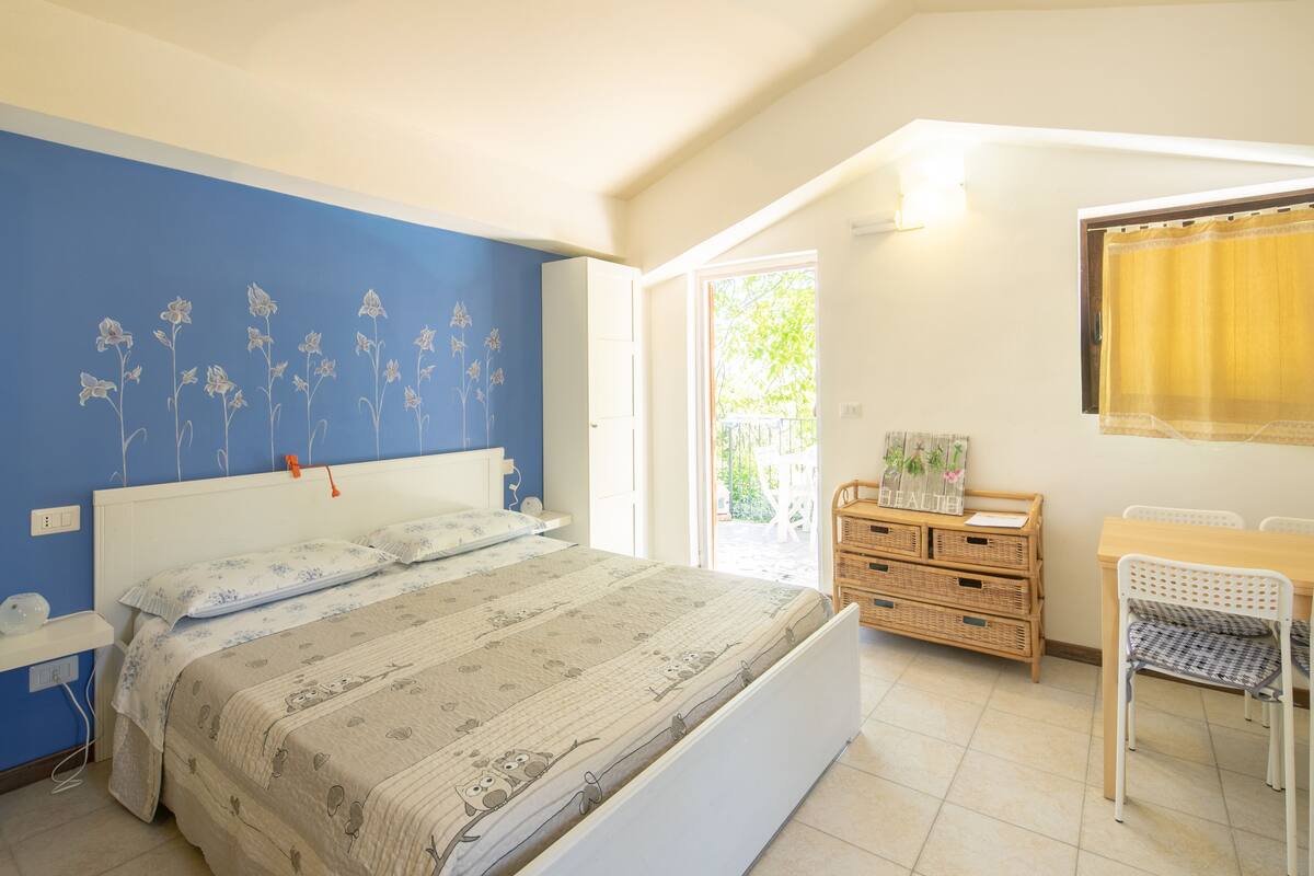 Een slaapkamer met een bed, een bureau en een stoel. Er is een raam met gordijn, een muur met bloemenmotief en een deur naar een buitenruimte.
