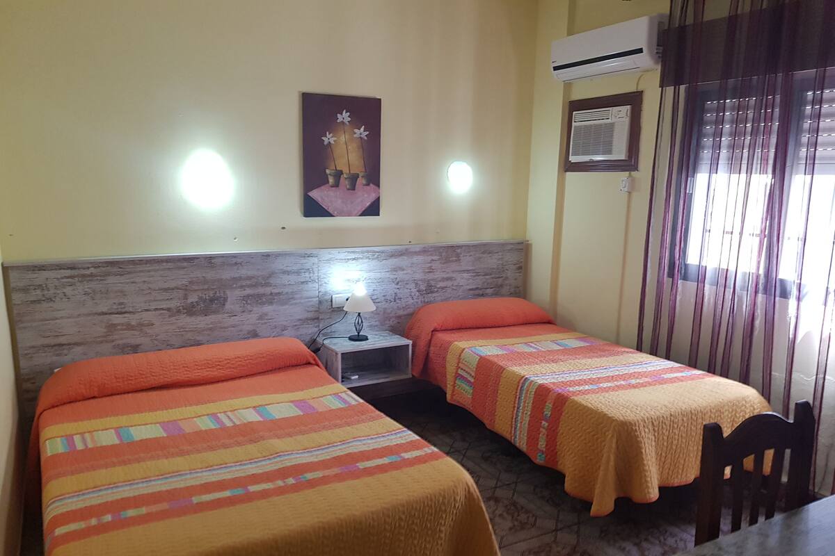Ein Hotelzimmer mit zwei Betten, einem Nachttisch mit Lampe, einem Bild an der Wand, einer Klimaanlage und einem Fenster mit Vorhängen.
