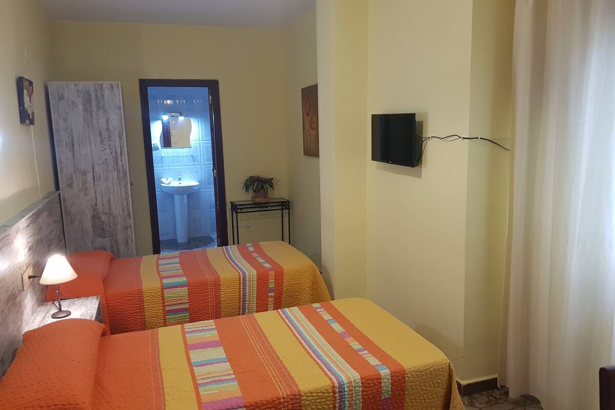 Ein Hotelzimmer mit zwei Betten, einem Nachttisch mit Lampe, einem an der Wand montierten Fernseher und einem durch eine offene Tür einsehbaren Badezimmer.