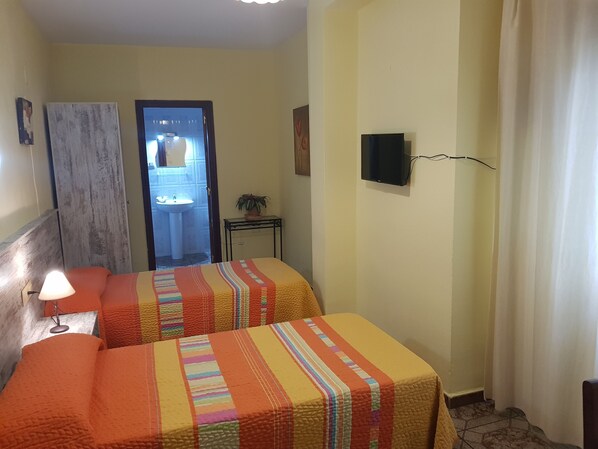 Een hotelkamer met twee bedden, een nachtkastje met lamp, een aan de muur bevestigde televisie en een zichtbare badkamer door een open deur.