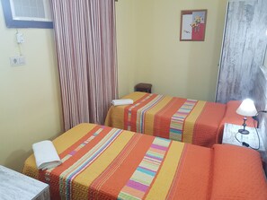 Een hotelkamer met twee bedden, gestreept beddengoed, een tafeltje met een lamp, een gordijn en een wandcontactdoos.