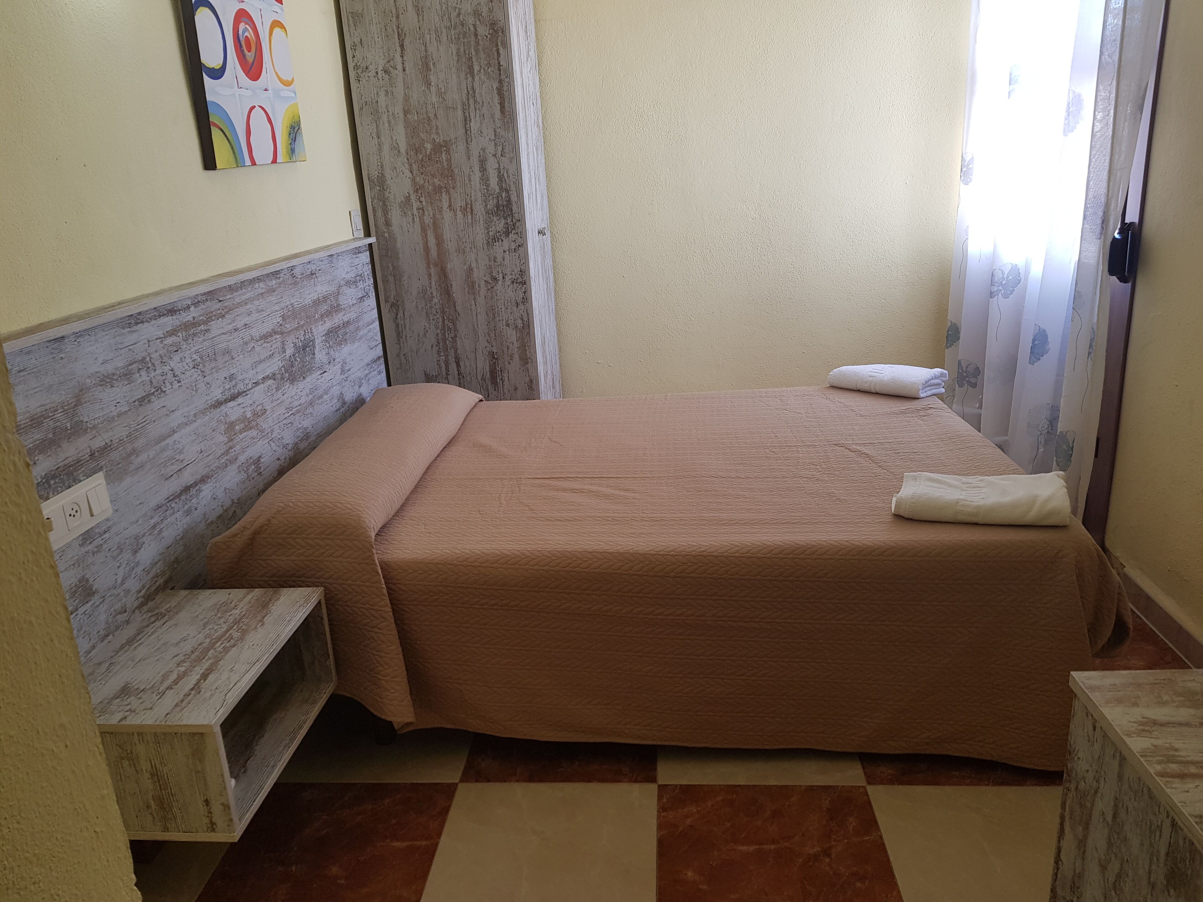 Un dormitorio pequeño y ordenado con cabecera de madera, una cama individual con colcha marrón, una mesita de noche y una obra de arte colgada en la pared.