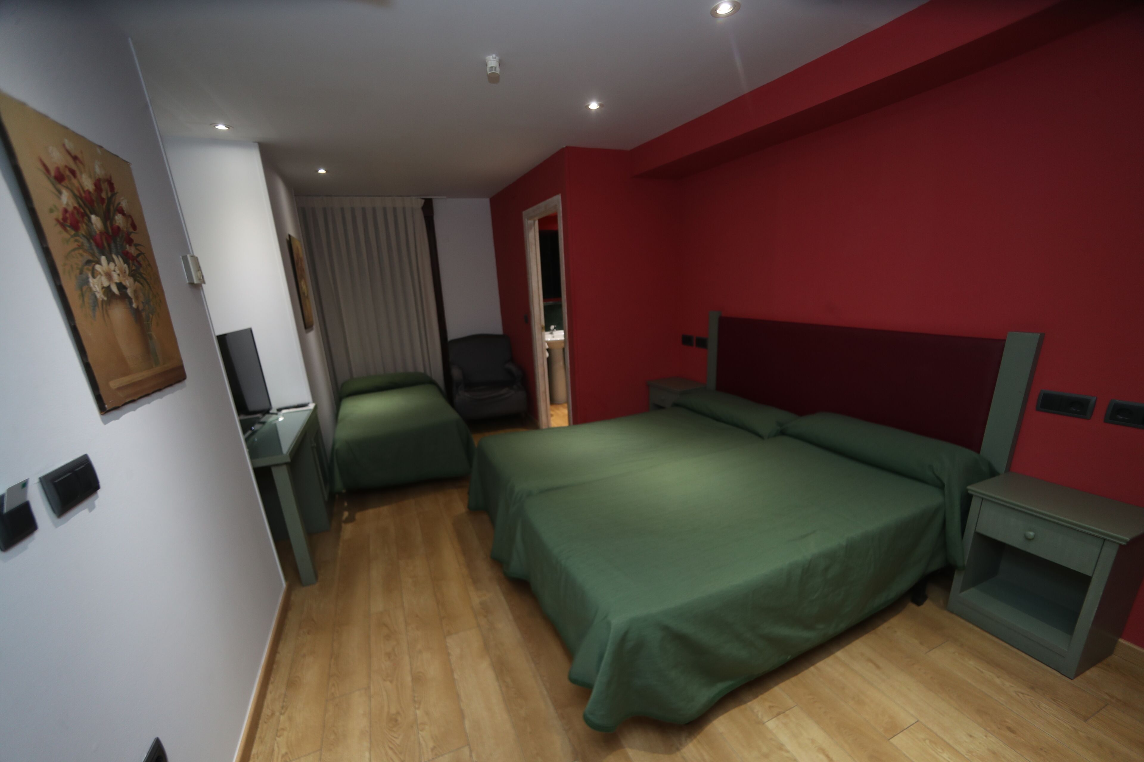 A hotel room with a green bed, a desk, and a chair. There is a painting on the wall and a window with curtains.