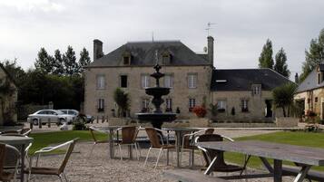 Un bâtiment en pierre avec une fontaine à l’avant, des places assises à l’extérieur et des voitures garées.