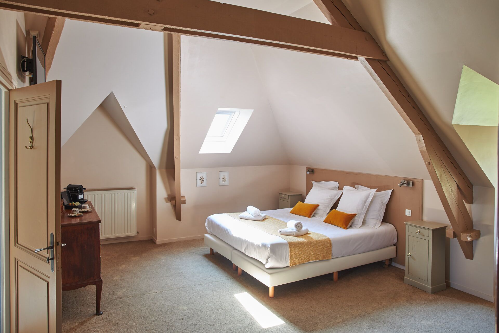 Ein Schlafzimmer mit schrägen Wänden, einem Bett mit weißer Bettwäsche und orangefarbenen Kissen, einer Holzkommode und einem Heizkörper.