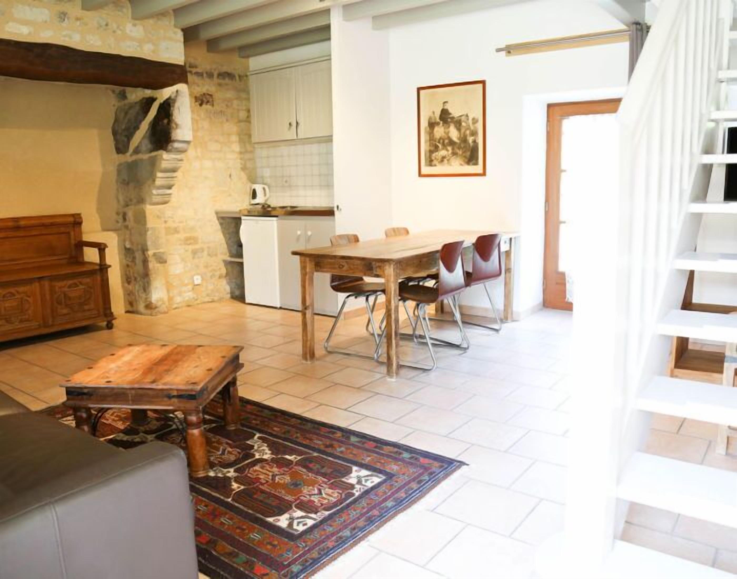 Amplia sala de estar con una mesa y sillas de madera, un sofá y un tapete estampado. Se ve una cocina con armarios blancos y una pared de piedra al fondo.