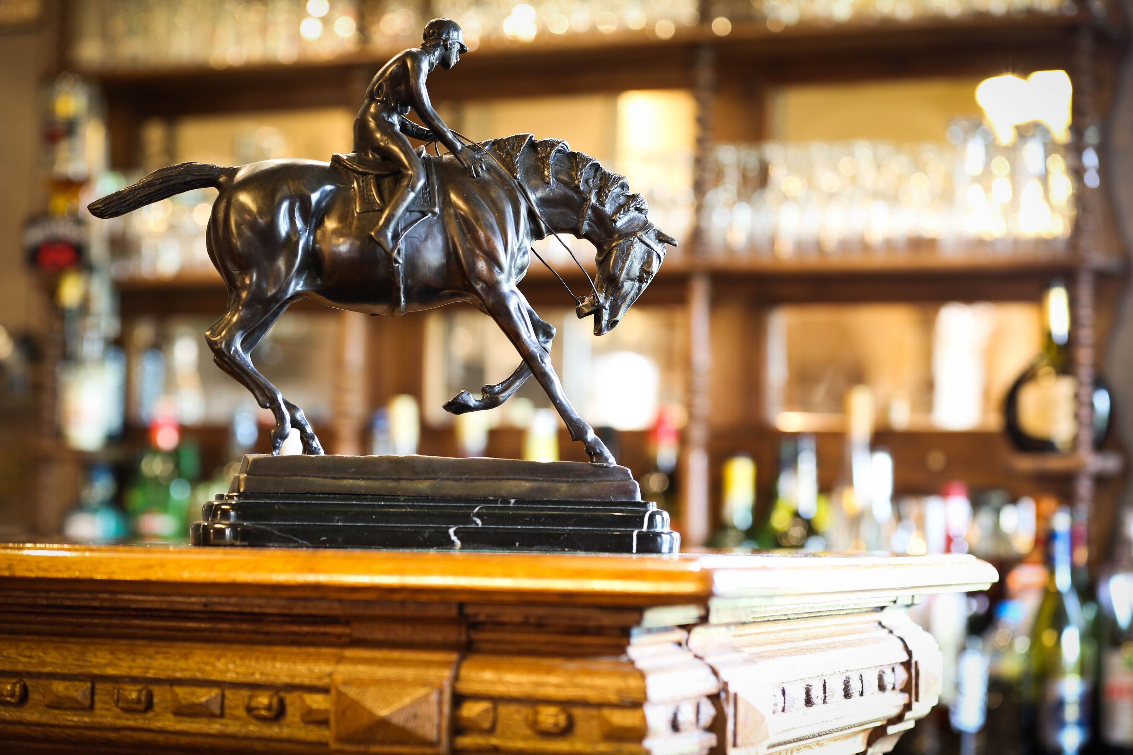 Angolo bar con una statua in bronzo di un uomo a cavallo, un bancone in legno e scaffali pieni di bottiglie sullo sfondo.