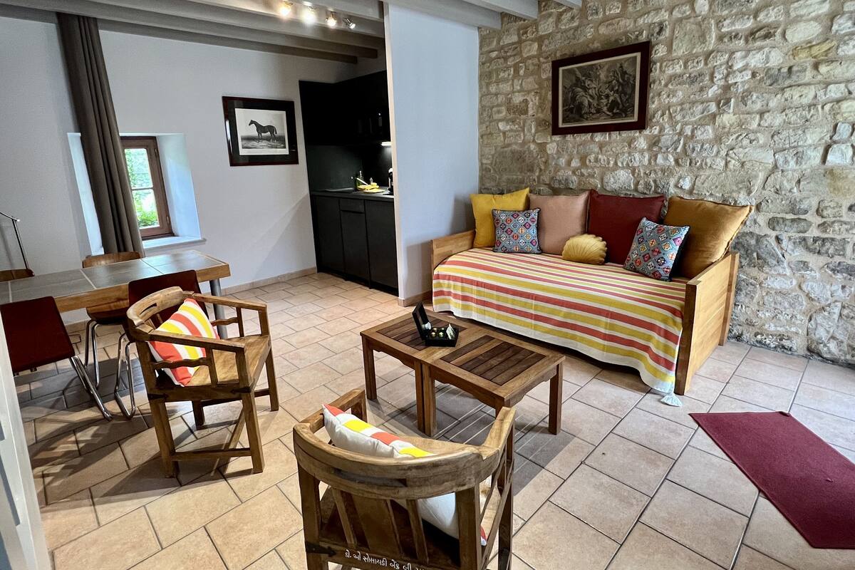 Un salon chaleureux avec un mur en pierre, un canapé en bois agrémenté de coussins colorés, une table à manger avec des chaises, et un coin cuisine à l’arrière-plan.