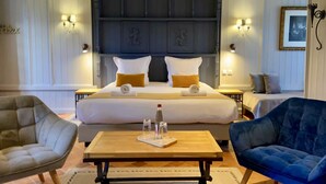Una habitación de hotel con una cama grande, dos mesitas de noche, una mesa de centro de madera, dos sillones y un cuadro enmarcado en la pared.