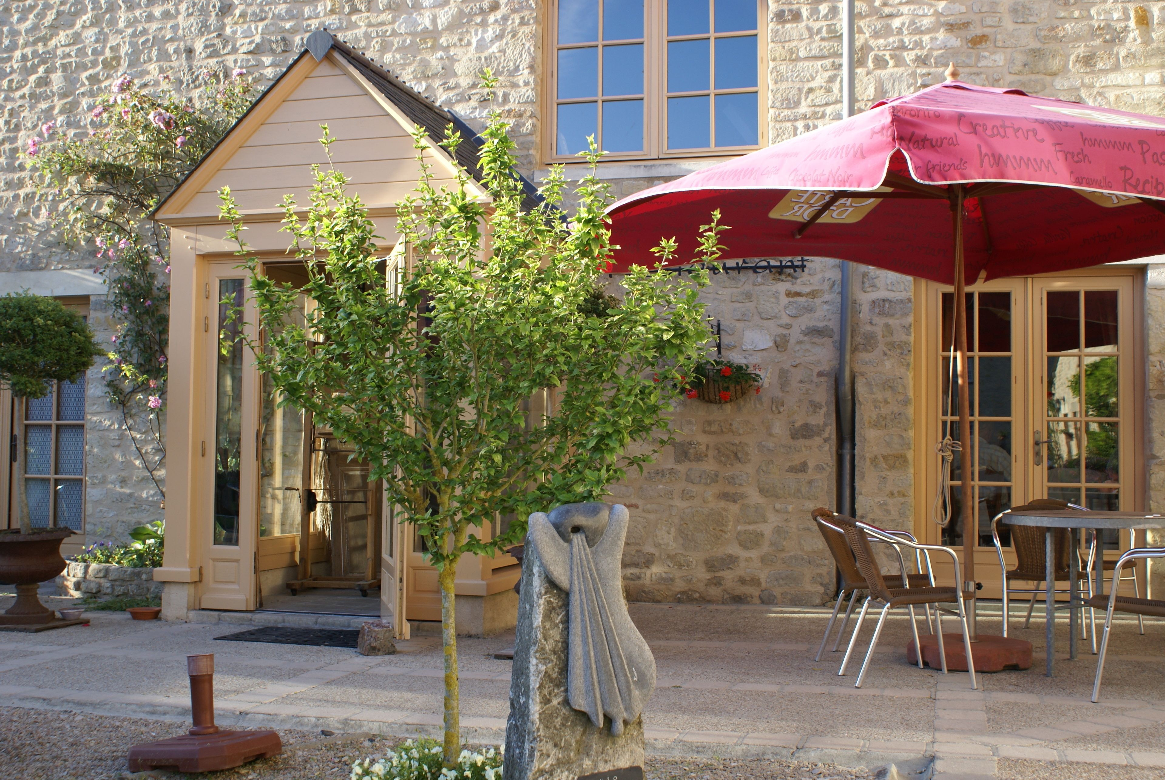 Un bâtiment en pierre avec une porte, un petit arbre, des sièges extérieurs avec des chaises en métal et un parasol rouge.