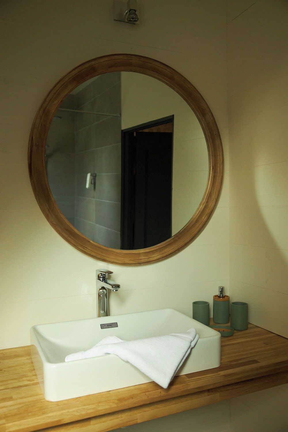 Ein Badezimmer mit rundem Spiegel, Waschbecken in Weiß und einem Holzlagerfläschchen.
