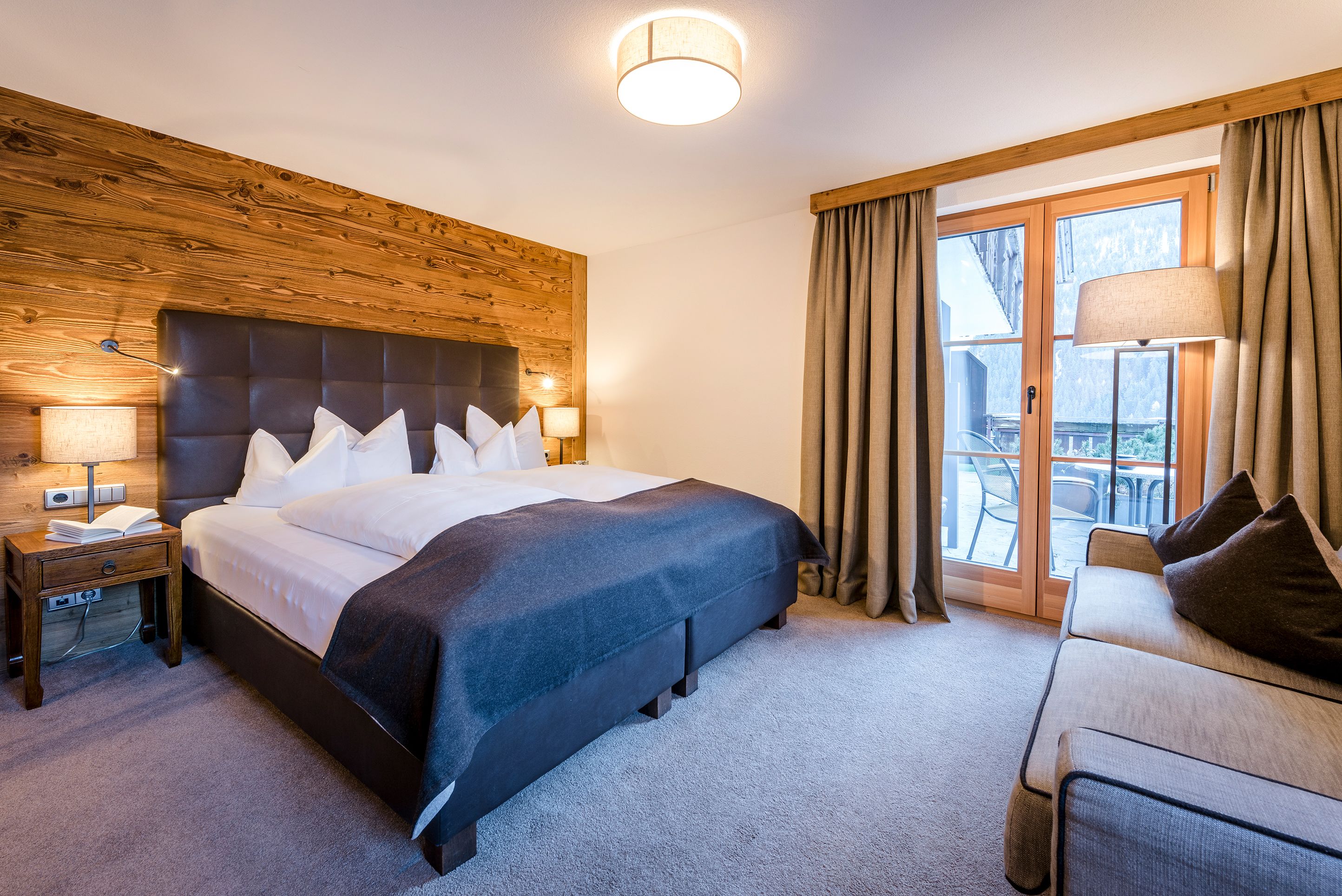 A modern hotel room with a large bed, a wooden headboard, a nightstand with a lamp, a sofa, and a view of the outdoors through a sliding glass door.