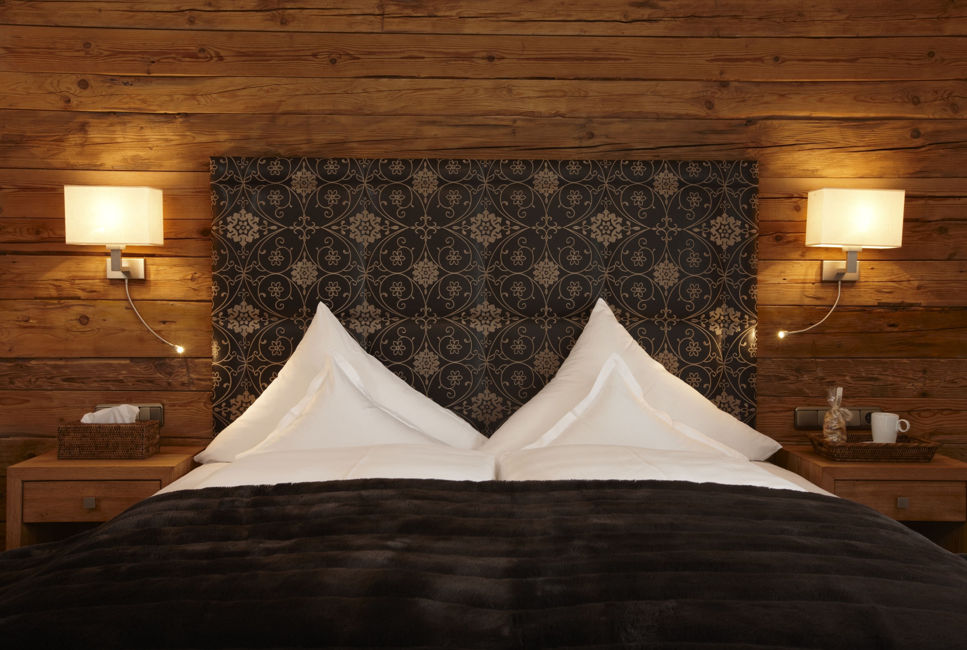 A bedroom with a wooden headboard, two bedside lamps, a dark patterned headboard, and a black bedspread.