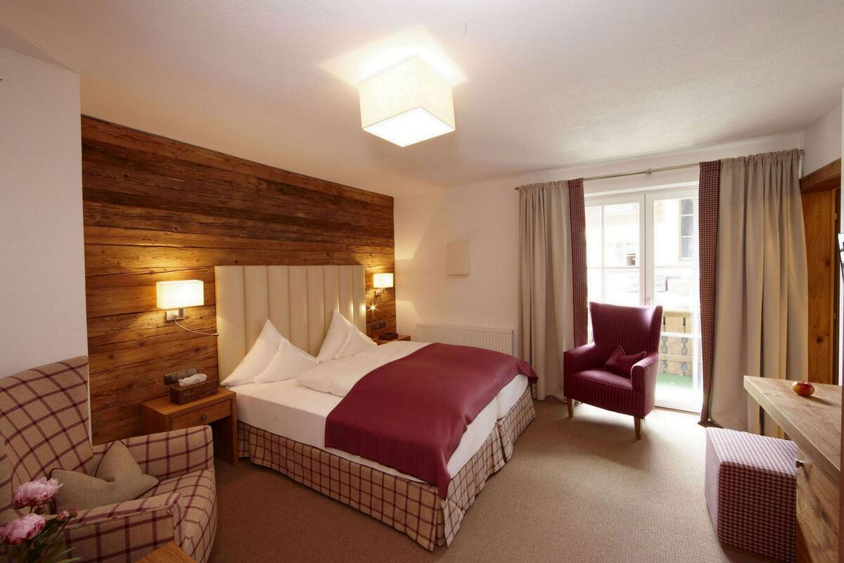 A hotel room with a wooden headboard, a bed with white linens and a red blanket, a plaid sofa, a chair, and a small table with a vase of flowers.