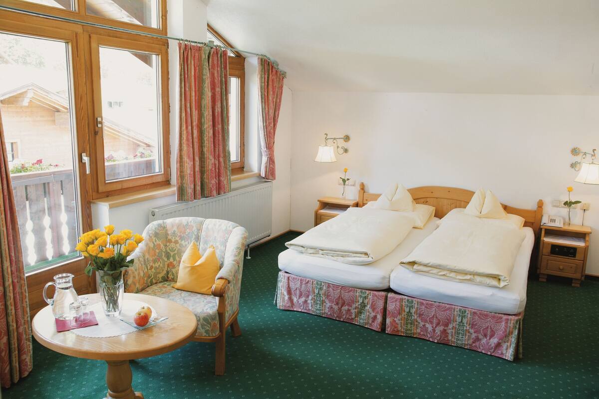 A room with a bed, a chair, a table with a vase of flowers, and a window with curtains.