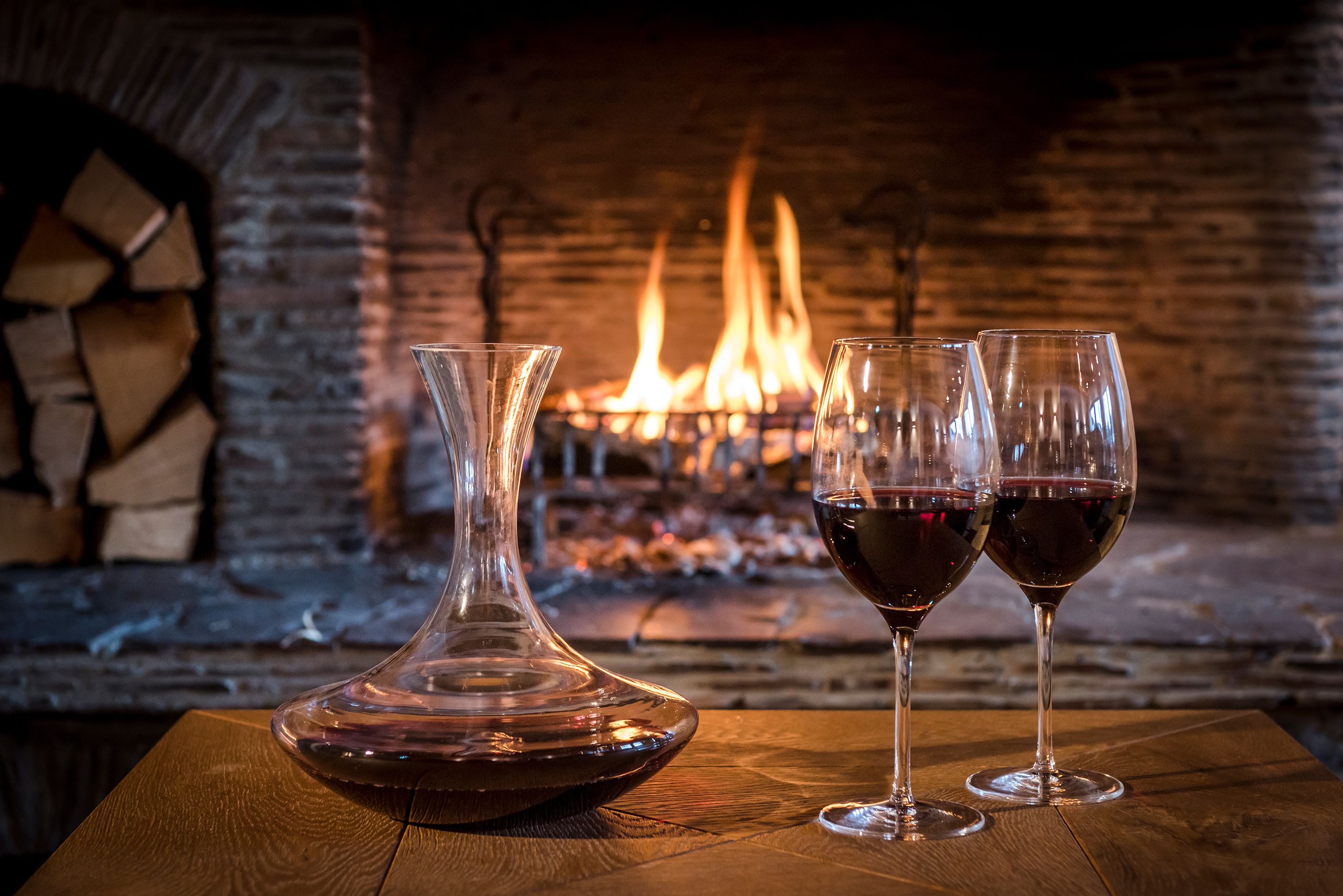 A fireplace with logs, two wine glasses, and a decanter on a wooden table.