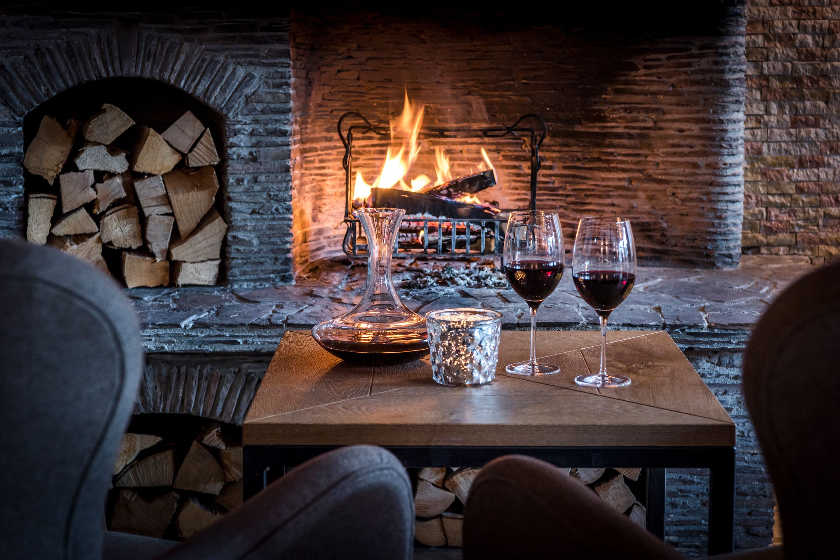 Un rincón acogedor con una chimenea, una mesa con vasos y una vela, y una pila de leña.