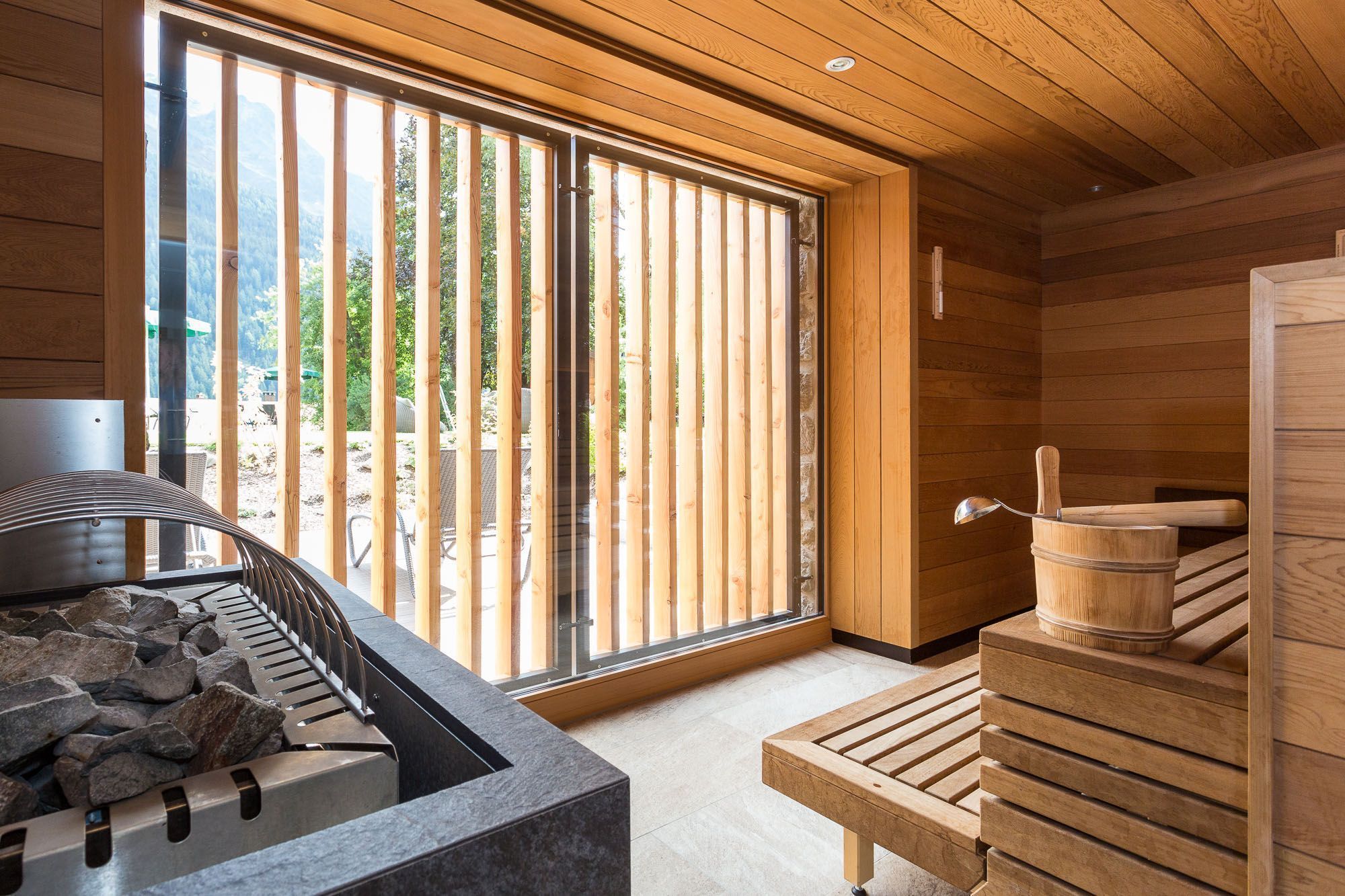 A sauna room with wooden walls, a stone bench, a sauna tub, and a view of the outdoors through large windows.