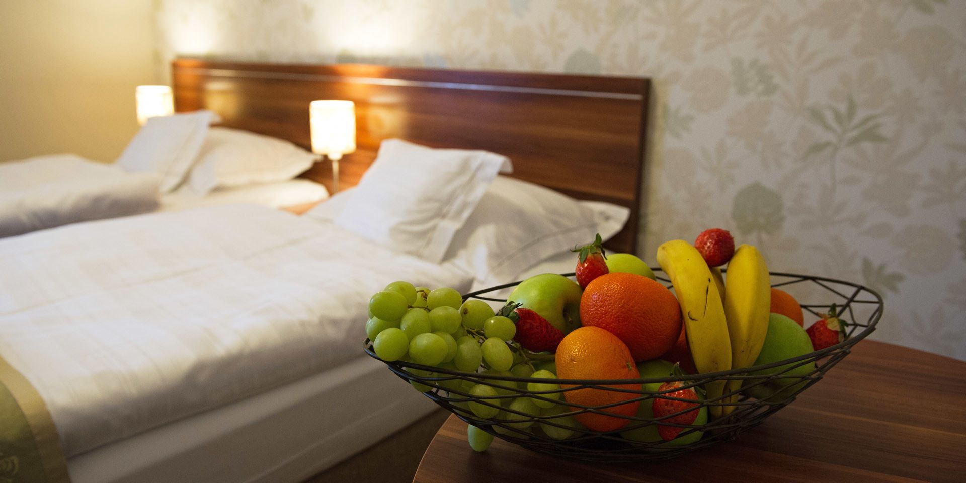 Habitación de hotel con dos camas, una mesita de noche con un frutero que contiene uvas, fresas, naranjas y plátanos.