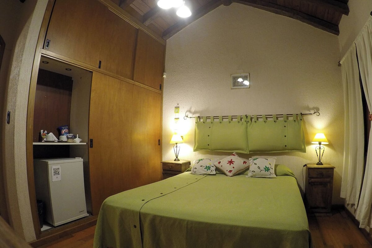 Une petite chambre à coucher accueillante, avec un lit simple, deux lampes de chevet, un petit réfrigérateur et une armoire en bois.