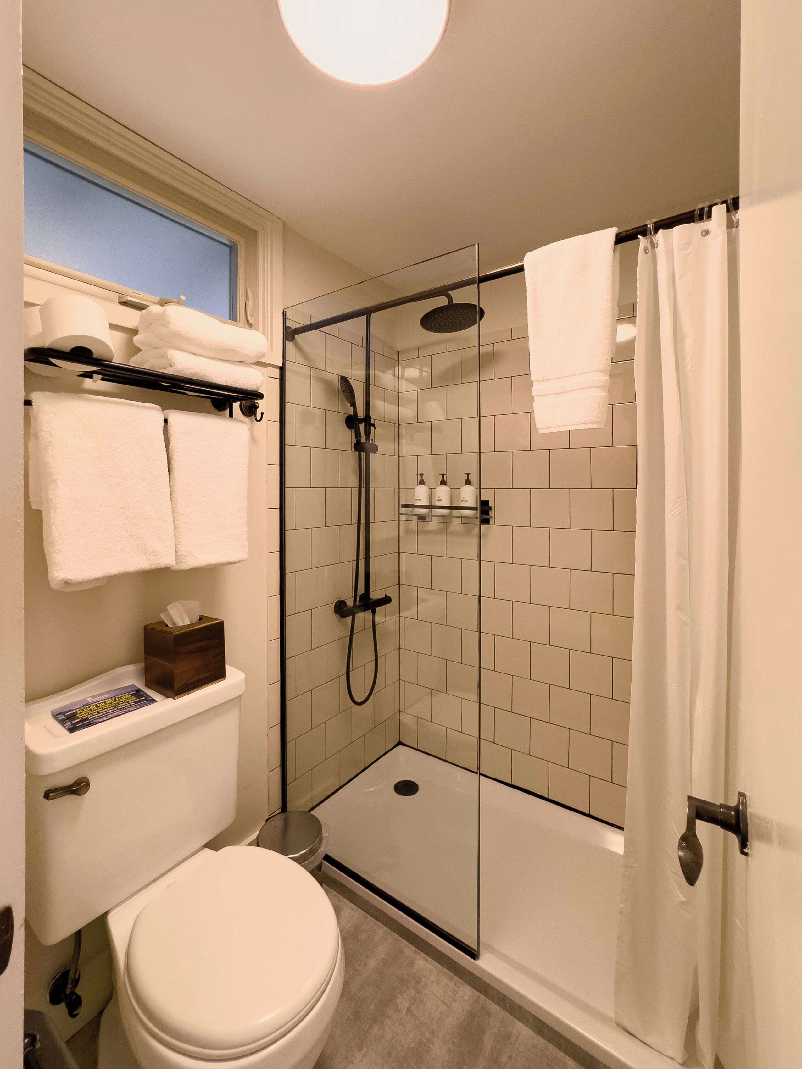 A modern bathroom with a glass-enclosed shower, white tiles, a toilet, and a window above the sink.