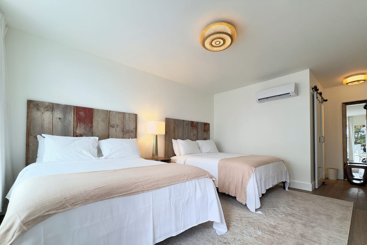 Hotelkamer met twee bedden, een houten hoofdeinde, een vloerlamp, een airconditioningsunit en uitzicht op de omgeving.