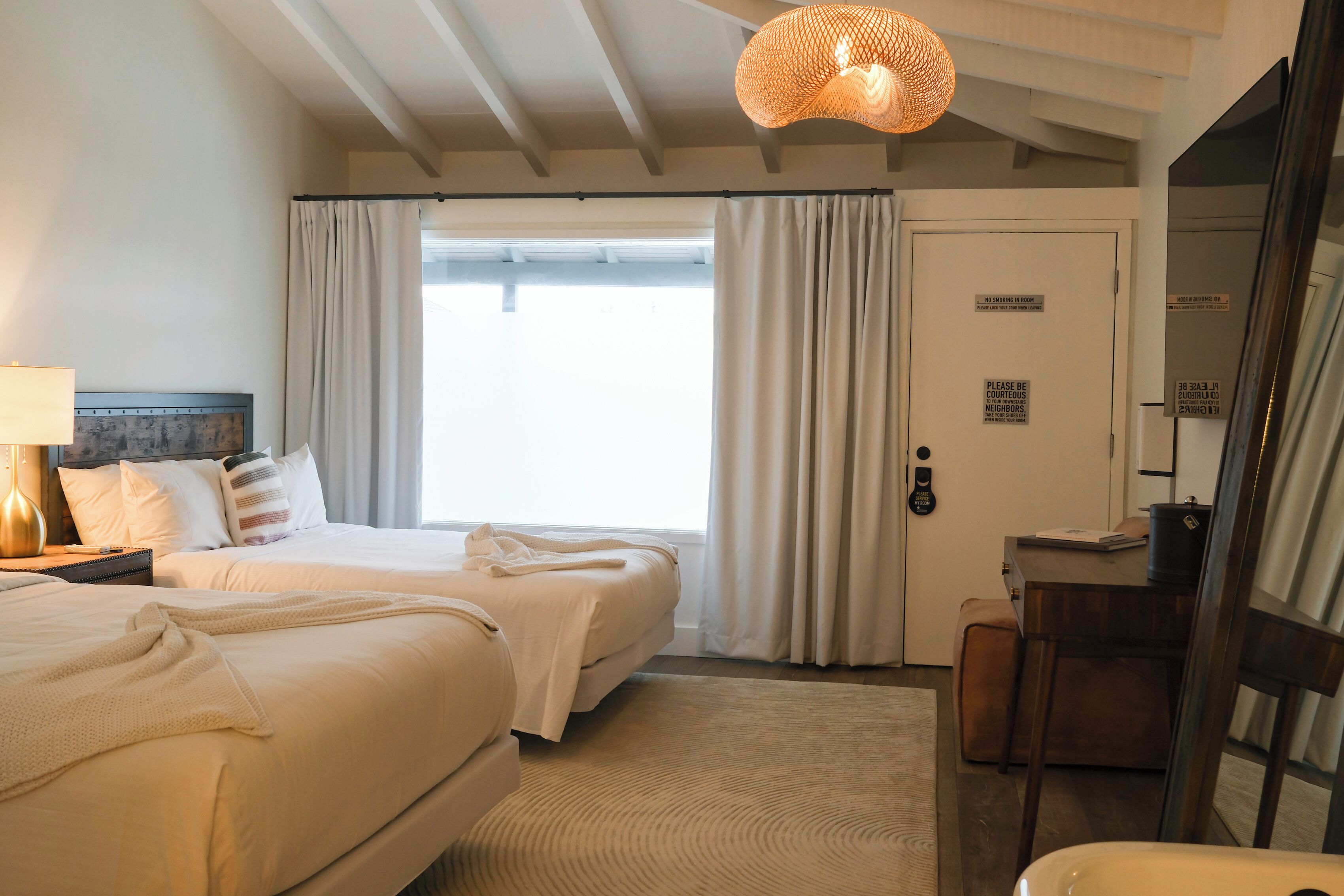 A hotel room with two beds, a desk, a chair, a lamp, and a large window with curtains.