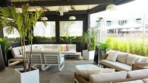 A covered outdoor seating area with white sofas and a dining table, surrounded by potted plants and large windows.